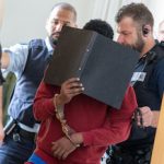The handcuffed defendant (C), covers his face with a file folder, as he is led by court officials into the hearing room of the Regional Court in Ulm. Photo: Stefan Puchner/dpa